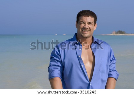 Muscular man in shirt smiling into camera.  Blue sky and ocean in the background.