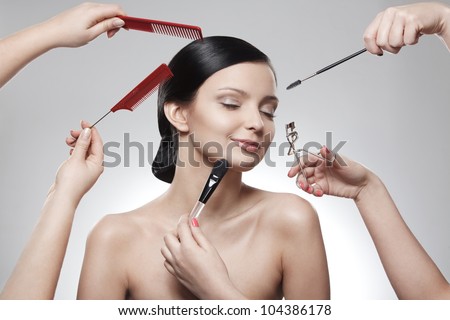 closeup portrait of happy girl and many hands with makeup brushes, eyelashes curling tongs and combs near her face over gray background