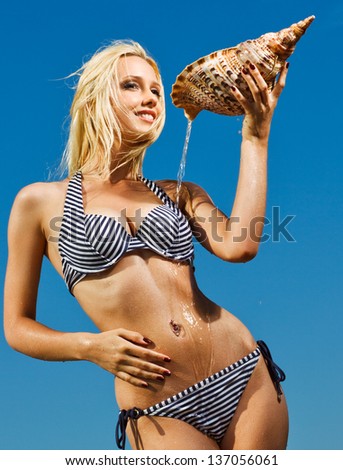 bright picture of happy sexy young woman pouring over herself water from the big seashell against blue sky