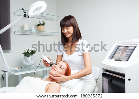 picture of pretty beautician doing rf-lifting procedure for young woman laying down in a beauty salon