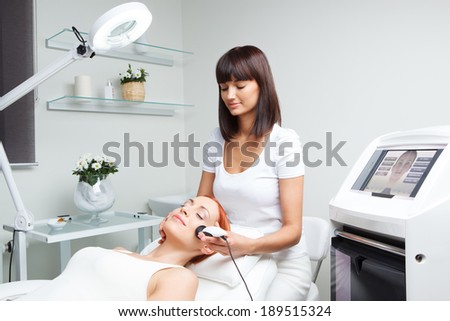 picture of pretty beautician doing rf-lifting procedure for young woman laying down in a beauty salon