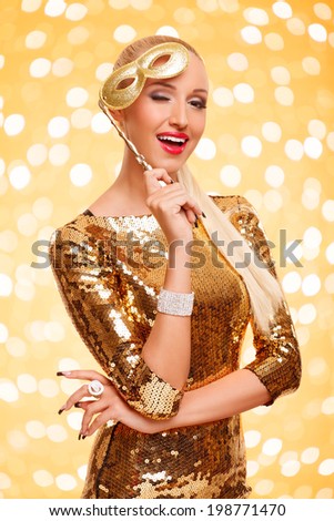 portrait of joyful winking girl in golden dress holding mask posing against gold background