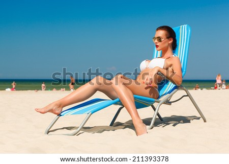 beautiful young woman in white bikini and sunglasses relaxing in deck chair on the sand beach