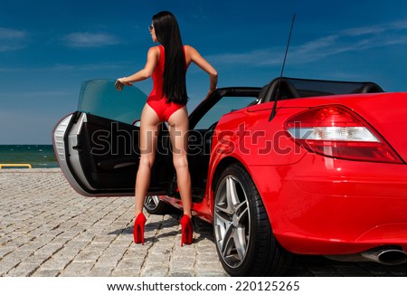 back view of sexy young woman in red bodysuit posing with red cabriolet against sea and sky background
