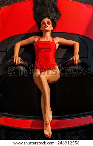 beautiful young woman with long dark hair in red bodysuit and sunglasses lying on the red cabrio