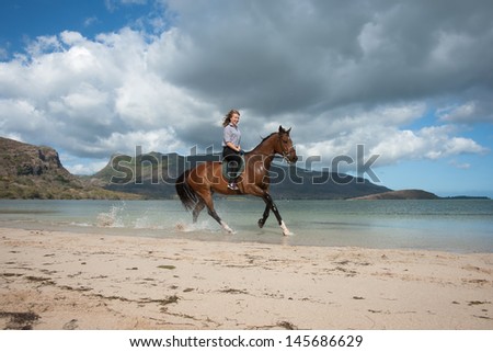 beautiful girl with long blond hair riding on a brown horse on the shore of Indian Ocean on the island of Mauritius