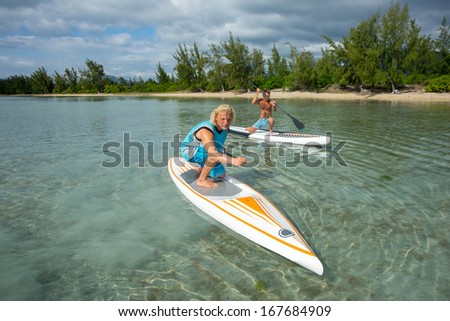 two young muscular surfers riding on the stand-up paddle board in the clear waters of the Indian Ocean  of Mauritius island on the background of green trees