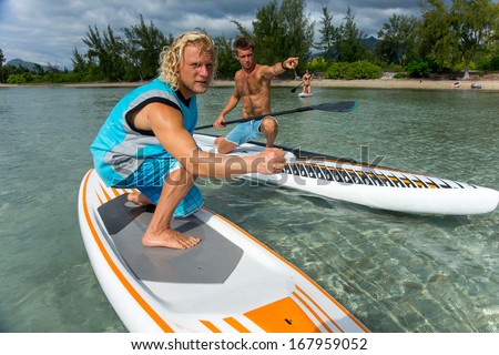 two young muscular surfers riding on the stand-up paddle board in the clear waters of the Indian Ocean  of Mauritius island on the background of green trees