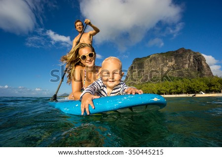 Young and beautiful parents ride by surfboard with baby in the Indian Ocean island of Mauritius on the background of mountains. They surf on a paddle board in transparent waters.