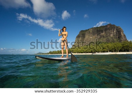 young beautiful girl-surfer riding on the stand-up paddle board in the clear waters of the Indian Ocean  of Mauritius island on the background of green trees and mountain