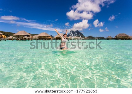 The girl jumps out of water lifting his hands up against the bungalow on stilts in the turquoise water from the sandy beach