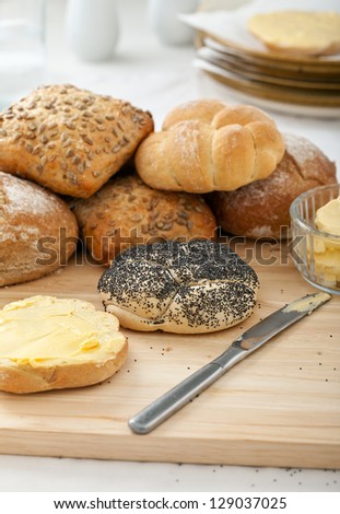 Mixed bread rolls stacked on bread board with butter spread on one roll shallow depth of field, kitchenware in the background.