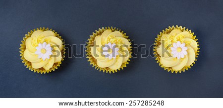 Aerial view of three lemon cupcakes with fondant flower in single file on a blue slate  background.