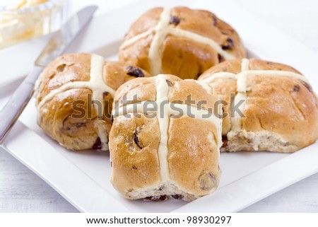 Hot Cross Buns. Traditional Easter spiced, sticky glazed fruit buns with crosses on white dish with butter dish and knife.