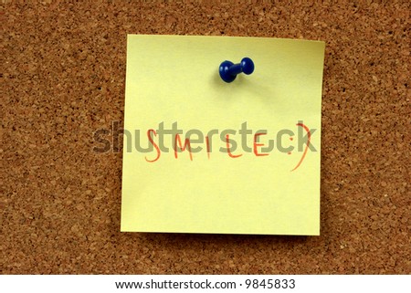 Yellow small sticky note on an office cork bulletin board. Smile!