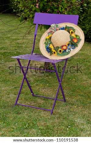 An old lilac folding chair with a broad-brimmed straw hat. On the hat there are some flowers.