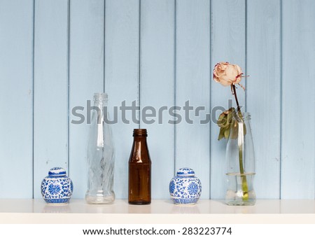 interior still life with bottles, rose and two china jars on the bookshelf on blue wooden background
