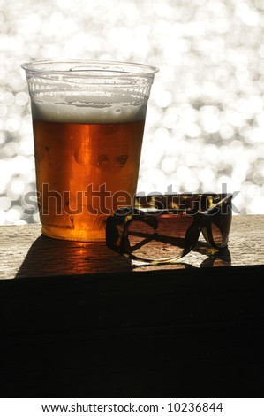 Glass of beer and a pair of sunglasses sitting on a deck handrail overlooking the ocean with space below for text