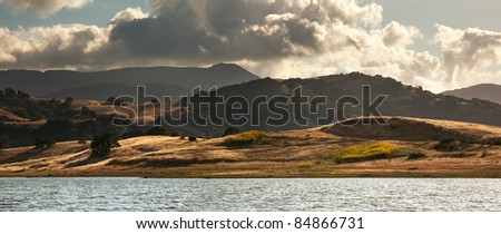 Panorama in of golden fields, mustard flowers, and the Calero Reservoir in summer in the Santa Cruz Mountains of California