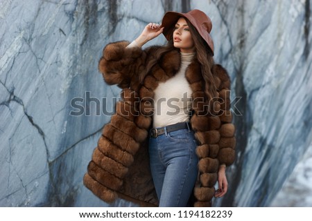 Young gorgeous woman with long wavy brunette hair in brown fur coat standing and posing against gray marble walls at stone quarry. Rich expensive woman