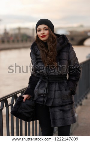Beautiful lady in gray fur coat and cashmere cap at river embankment on a cold day. Fall winter fashion look.