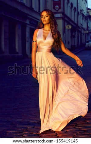 fashion portrait of young beautiful girl in pale lilac color long flying dress walking down the street in old town at sunset