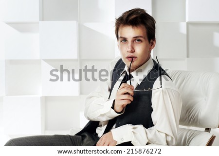 Young handsome stylish man sitting in armchair in modern interior holding his glasses and looking in camera