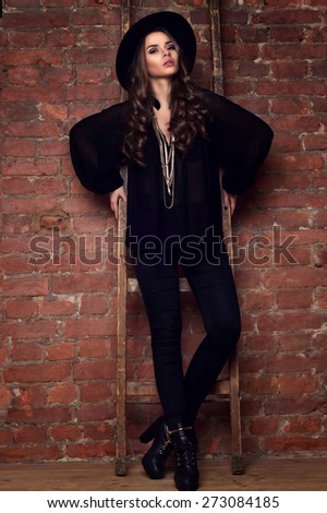 Fashion style portrait of young trendy girl wearing black clothes: blouse, trousers, hat and boots. Model posing and standing near brick wall and wooden ladder.
