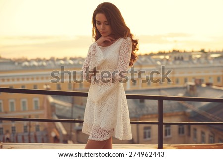 Summer outdoor porttrait of young pretty elegant girl posing at roof at sunset. Lovely soft back light. View on city buildings and roofs.