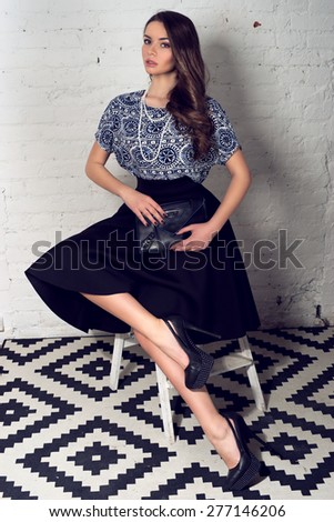 Fashion portrait of young pretty stylish girl in black skirt and blue blouse sitting at small staircase and holding black leather handbag
