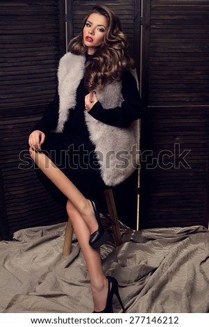Fashion portrait of young lady in black dress and fur coat sitting on a stool. Pretty girl with dark long curly hair.