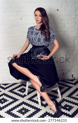 Fashion portrait of young pretty stylish girl in black skirt and blue blouse sitting at small staircase and holding black leather handbag