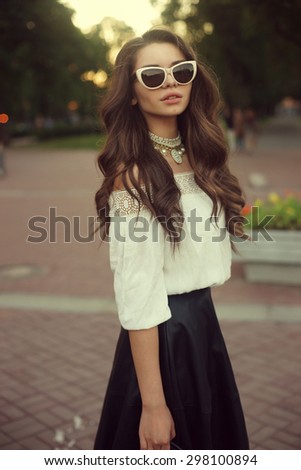 Outdoor summer portrait of young stylish classy woman with long curly brown hair. Elegant girl posing in white blouse, black skirt,sunglasses and necklace in city park at sunset. Soft light full body