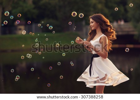 Outdoor summer portrait of young beautiful happy woman making soap bubbles in park or at nature. Joyous happy girl in white dress