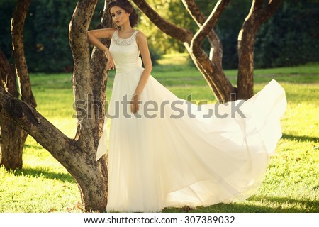 Wedding. Young beautiful bride in white flying dress standing near tree in park at sunset. Soft lightning