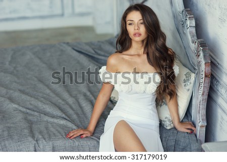 Young beautiful and sexy woman sitting on bed in white luxury evening dress and dreaming or thinking. Fashion style portrait of sexy girl with long curly hair