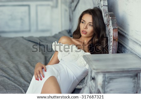 Beautiful stunning woman wearing white evening dress and sitting or lying on the bed in a luxury interior. Fashion style portrait of pretty lady