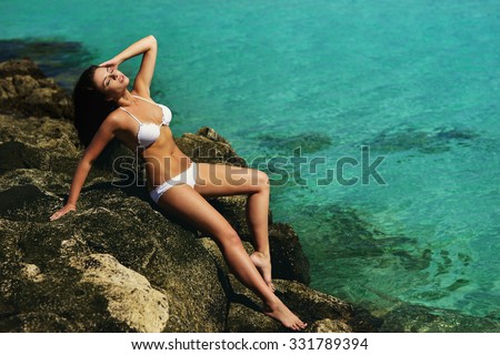 Fashion style full length portrait of young beautiful sexy tanned girl in white bikini sitting at rocks near blue green clear ocean sea water. Sunbathing woman. Vogue style