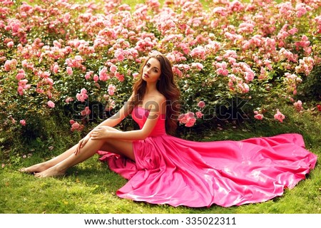 Young beautiful pretty girl with curly hair in pink silk dress sitting on grass near bushes of roses in blossom