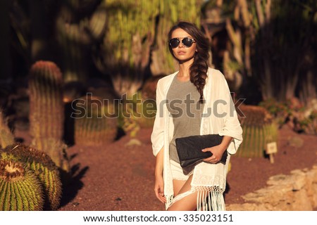 Stylish trendy beautiful girl with long braid posing in t-shirt and shorts with handbag against cactuses. Outdoor summer portrait