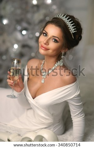 Elegant rich lady wearing white evening dress and jewelry with diamonds sitting on bed in christmas decorated interior and drinking champagne. Beautiful smiling woman looking in camera