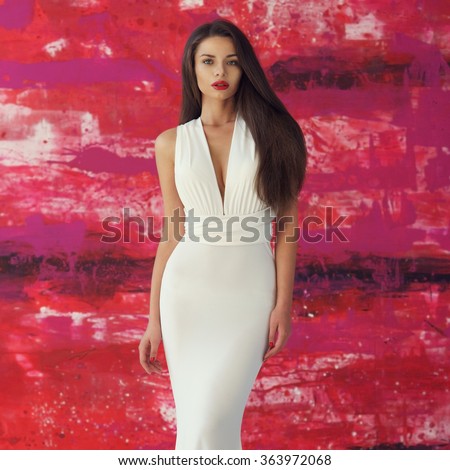 Young beautiful stunning woman posing in long elegant white evening dress and red shoes against stylish red background