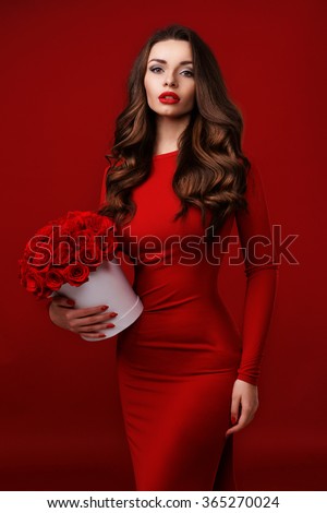 Young beautiful pretty girl standing and holding white box with red roses. Vogue fashion style studio portrait of sexy girl in red dress