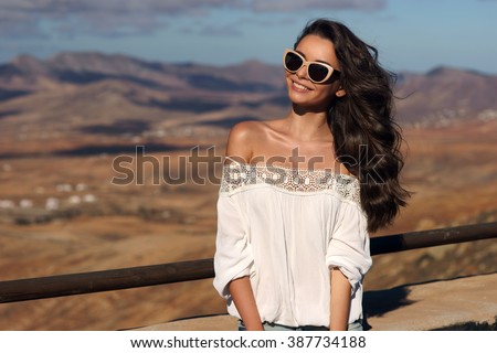 Young beautiful stylish happy smiling girl standing against magnificent view on valley and hills at sunset. Pretty woman traveling