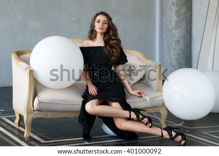 Elegant calm woman in black dress and shoes with long brunette curly hair sitting on sofa in interior in minimalism style and holding white balloons. Vogue fashion style portrait