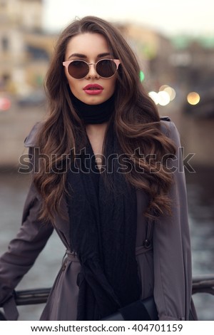 Fashionable glamorous girl with long curly hair wearing black dress, scarf, classy coat and sunglasses posing at city streets and looking at you or in camera. Fashion vogue style outdoor portrait