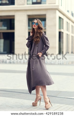 Outdoor dynamic fashion portrait of young beautiful stylish woman in black dress, grey coat and sunglasses walking on a windy day against city background
