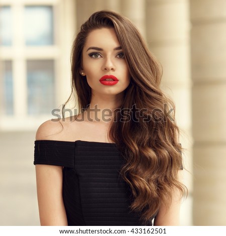 Close up portrait of young beautiful woman with long brunette curly hair posing against architectural background and looking at you