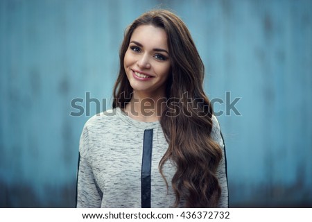 Happy attractive smiling girl portrait against blue old wooden wall. Pretty stylish fashionable woman in gray hoody with long curly hair looking at you. Shallow DOF, blurred background