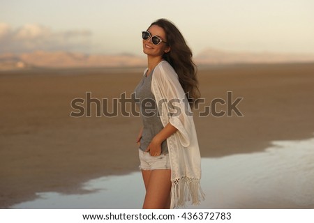 Young beautiful girl walking at the beach at sunset. Woman in gray t-shirt, white cape and shorts posing enjoy sunset